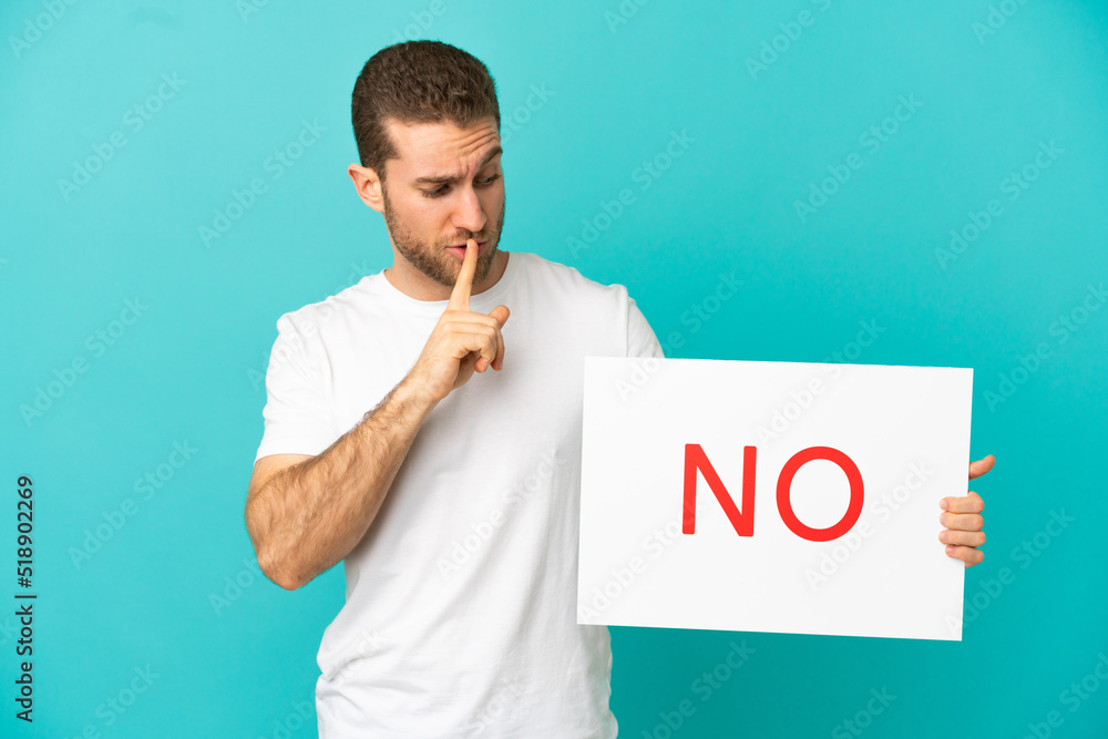 Handsome blonde man over isolated blue background holding a placard with text NO doing silence gesture Handsome blonde man over isolated blue background holding a placard with text NO doing silence gesture