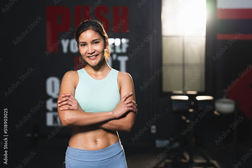 Smiling Asian athlete woman in sportswear standing with arm crossed posing after exercise in gym.Portrait female workout bodybuilder with sweat showing abdominal muscles at sport club fitness.