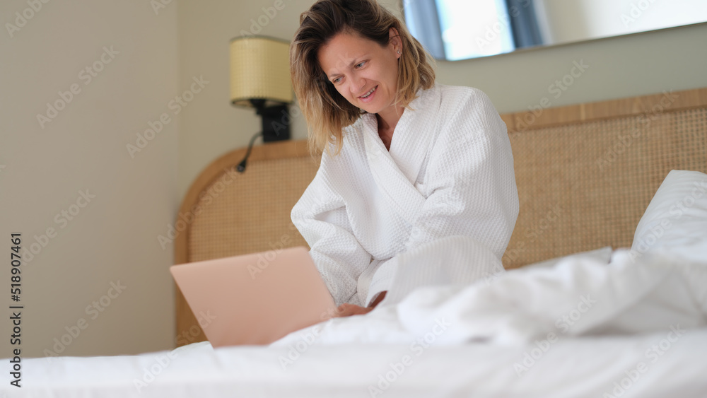 Woman sits at home on bed in white bathrobe and works on laptop Woman sits at home on bed in white bathrobe and works on laptop
