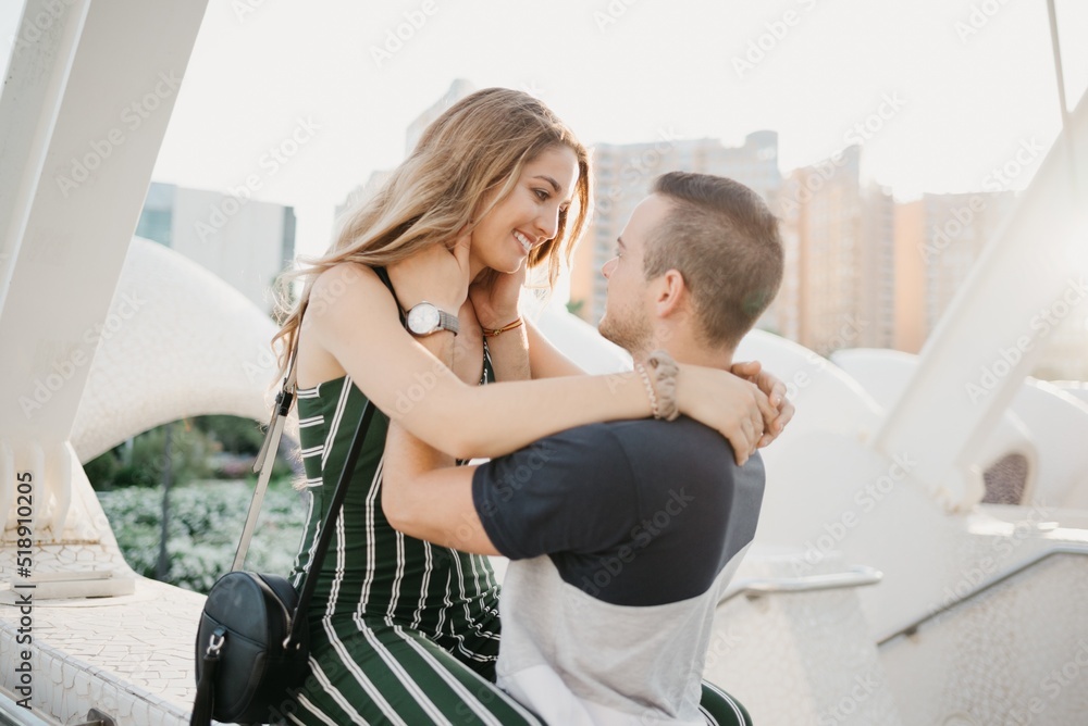 A close photo of a smiling girl who is hugging her boyfriend in the modern urban space at sunset in Spain. A couple of tourists on a date in the evening in Valencia.