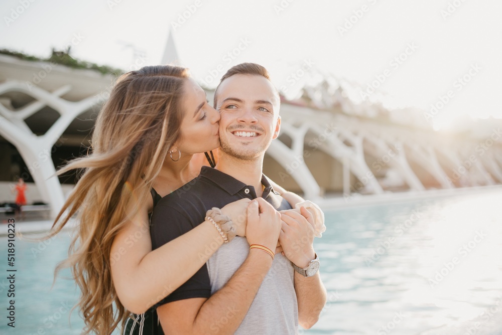 A smiling girl is kissing her boyfriend from the back in the modern urban space at the sunset in Spain. A couple of tourists on a date in the evening in Valencia.
