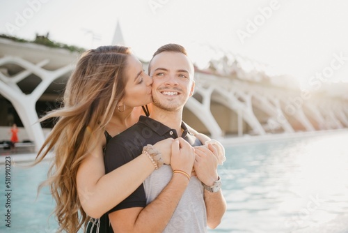 A smiling girl is kissing her boyfriend from the back in the modern urban space at the sunset in Spain. A couple of tourists on a date in the evening in Valencia.