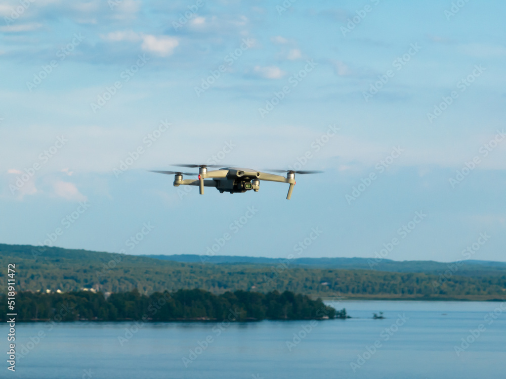 A consumer drone is seen in-flight, outdoors, high above a lake with a forest in the landscape background.