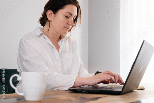 Middle age woman freelancer is working at home. Pretty plus size female in 40s working with laptop and phone from her kitchen in the morning with mug of hot drink. Real people lifestyle © ninelutsk