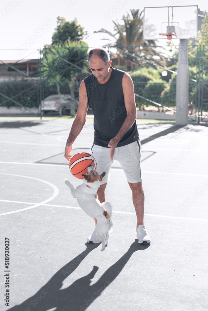 Portrait of adult senior man playing sport basketball ball with a dog jack russel terrier on playground outdoor. A pet jumping to catch the ball.