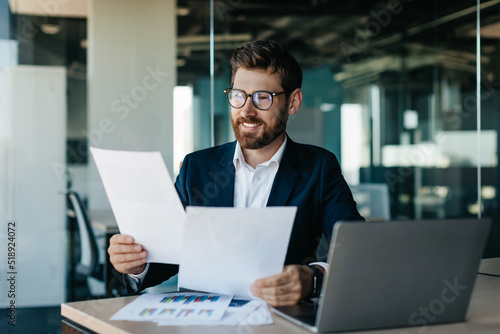Happy businessman reading papers and business reports, rejoicing in the affairs of company, sitting in modern office photo