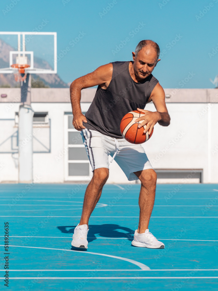 Adult senior man playing a sport ball on blue basketball court at summer sunny day. Adult senior man playing a sport ball on blue basketball court at summer sunny day.
