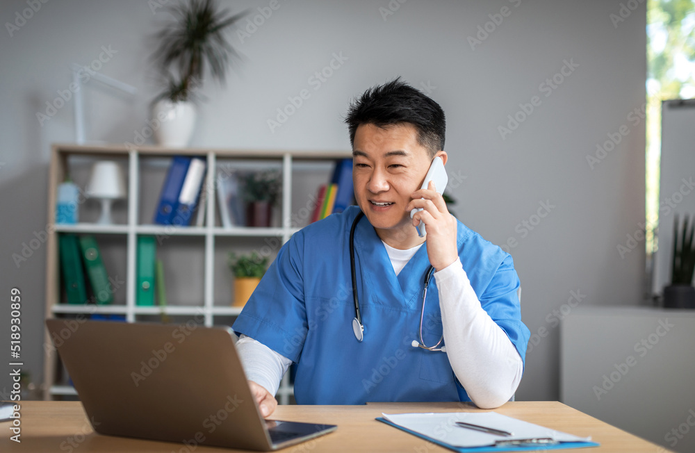 Smiling millennial asian man doctor in uniform call by phone, types on computer in office clinic interior Smiling millennial asian man doctor in uniform call by phone, types on computer in office clinic interior
