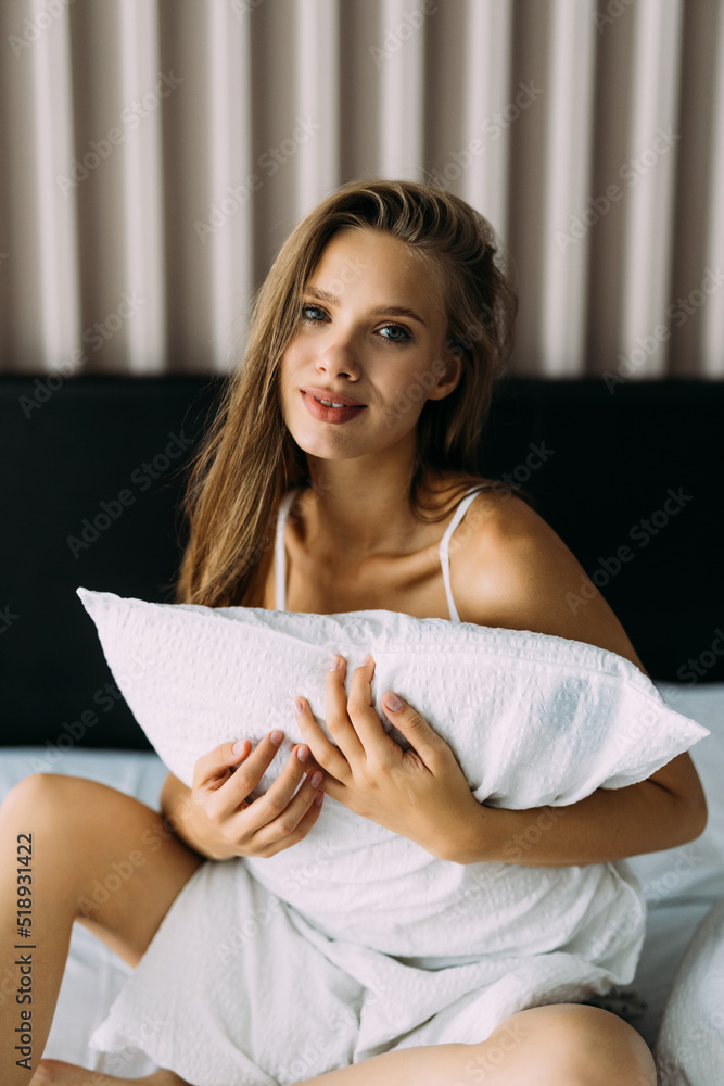 Attractive young woman sitting on the bed covering her body with a big white pillow Attractive young woman sitting on the bed covering her body with a big white pillow