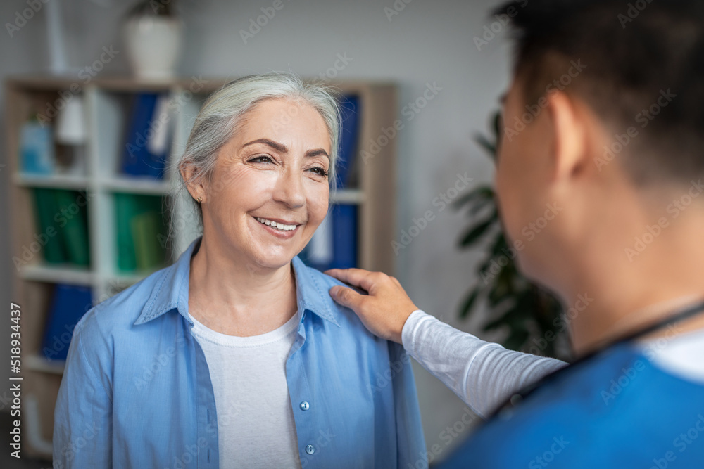 Cheerful senior european lady patient on meeting with young chinese doctor in uniform talking in office Cheerful senior european lady patient on meeting with young chinese doctor in uniform talking in office