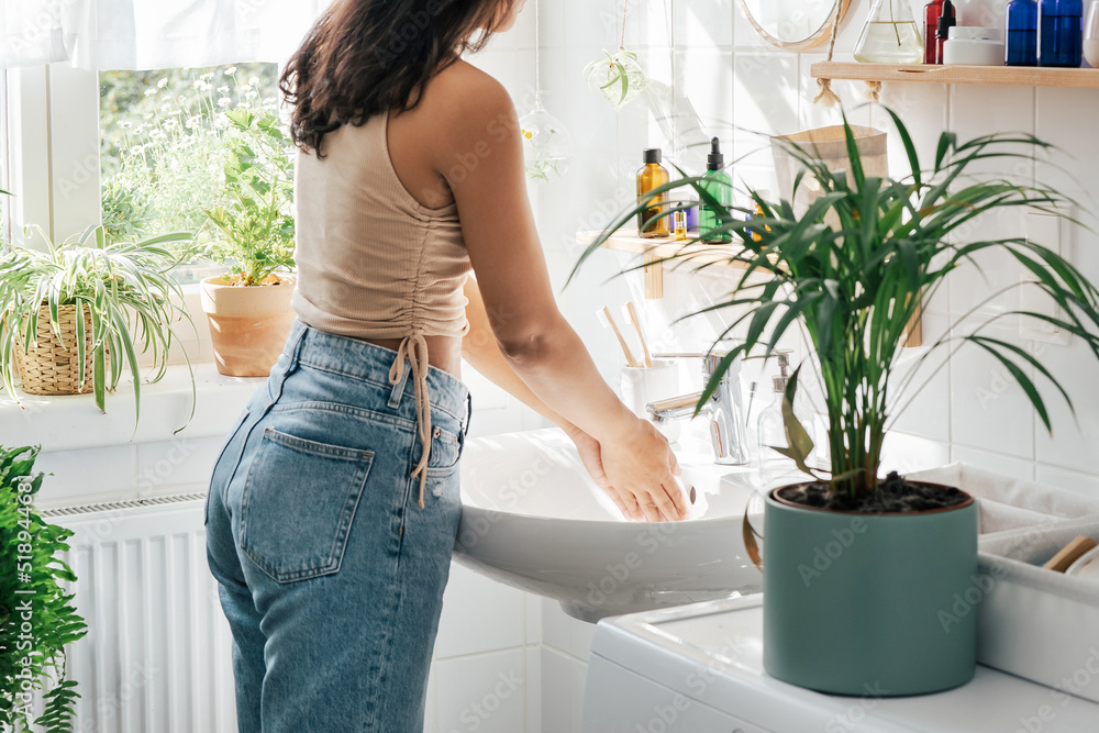 Anonymous young hispanic woman washing hands in white bathroom with green plants. Wellness concept