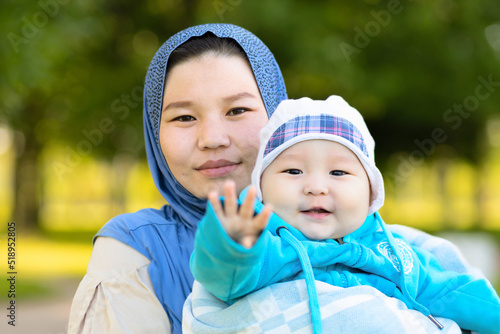 Cute young islamic kazakh woman hugging, playing with her little son, newborn boy. Yappy asian mother in hijab holding baby in hands outdoors. Family concept