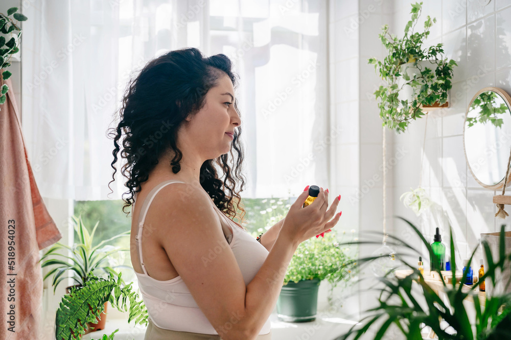Young brunette woman in pink underwear holding glass cosmetics bottle in white bathroom with biophilic design. Body positivity, natural cosmetics, home comfort zone Young brunette woman in pink underwear holding glass cosmetics bottle in white bathroom with biophilic design. Body positivity, natural cosmetics, home comfort zone