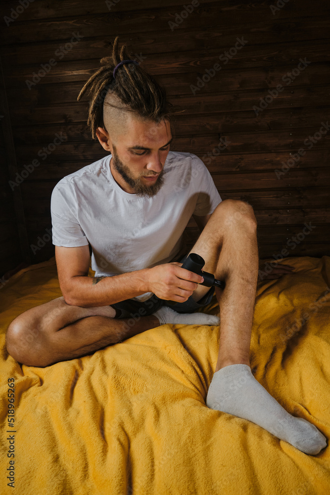 Caucasian man with dreadlocks gives himself massage with percussion gun at home sitting on bed. Athlete kneads leg muscles after training. The concept of healthy lifestyle.