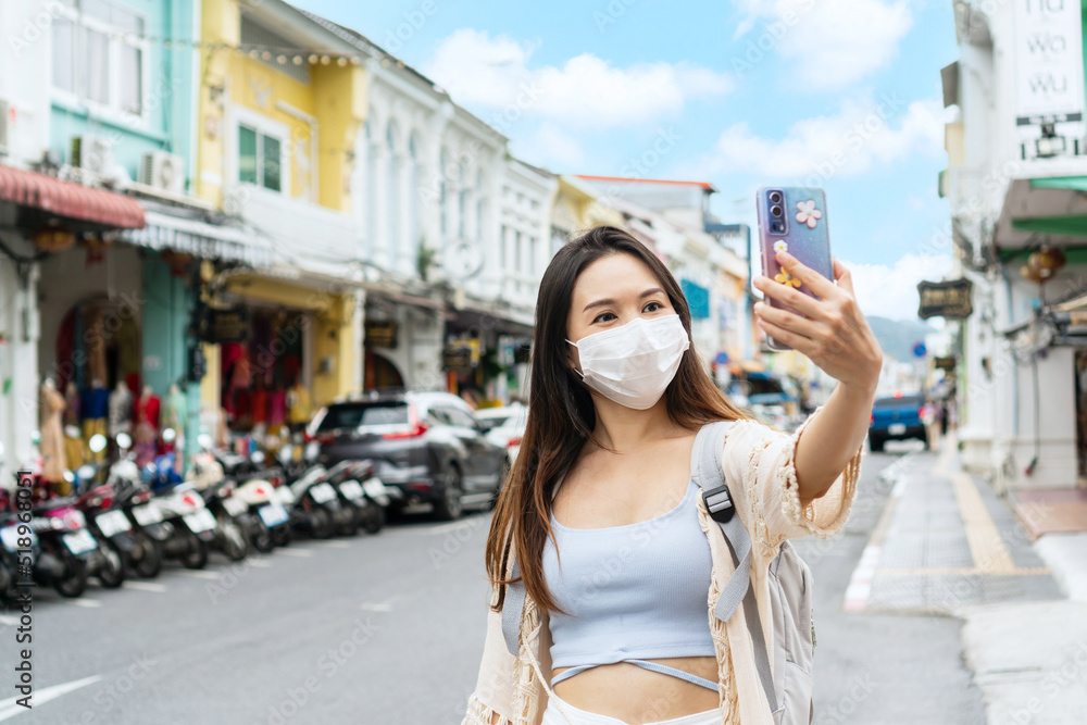 Asian traveler wearing a medical mask take a photo or video call with her family and smiles while walking in old town Phuket, Thailand. Travel during Covid-19 pandemic, new normal lifestyle concept