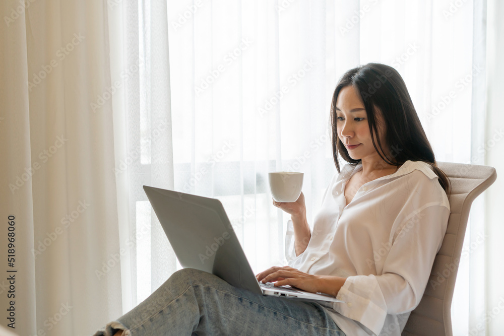 Happy Asian woman using laptop while holding a cup of coffee at home. Smiling female working on computer, enjoying internet shopping or chatting in social networks. Technology and lifestyle concept. Happy Asian woman using laptop while holding a cup of coffee at home. Smiling female working on computer, enjoying internet shopping or chatting in social networks. Technology and lifestyle concept.