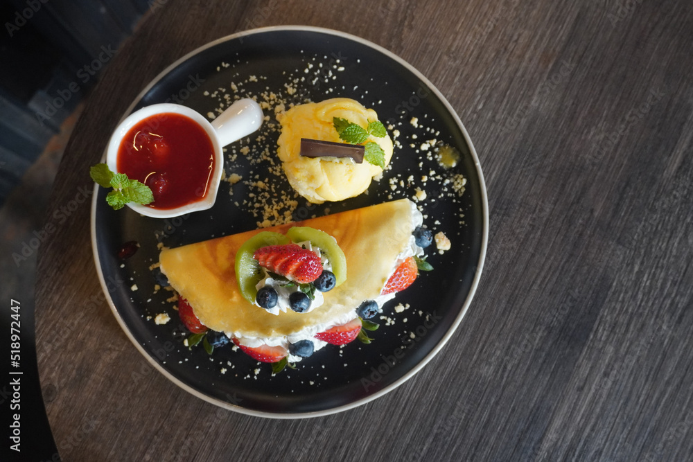 top view crispy crape and mix fruit with ice cream passion fruit, fresh strawberry fruit and strawberry Jam in a white ceramic cup on black ceramic plate, on wooden table background