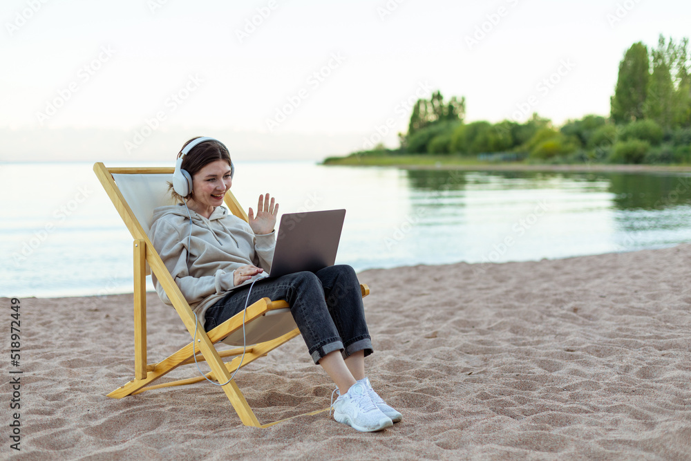 Woman in headphones video chatting by laptop, saying hello sitting on a beach