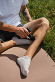 Concept of healthy lifestyle. Athlete stretches leg muscles after training using electronic body machine sitting on sports mat in park. Caucasian man massages himself with percussion gun outside.