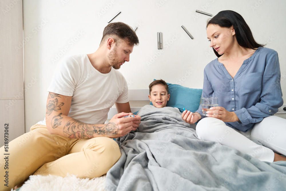 Parents atbedside of asick son with thermometer and pills Parents atbedside of asick son with thermometer and pills