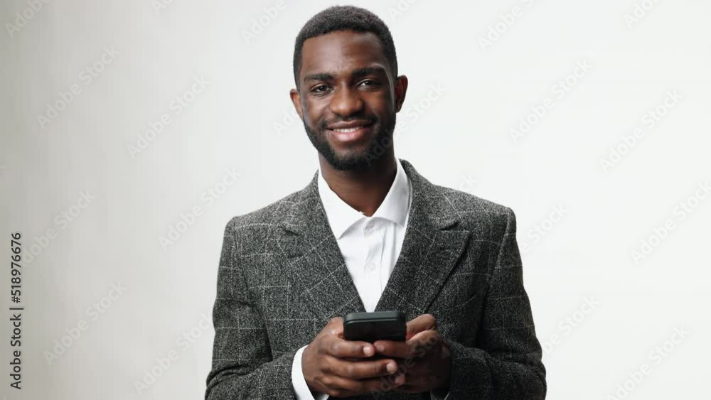 Man with phone in hand African-American man in business office business suit looking at phone and smiling and scratching his head with his hand on white background Man with phone in hand African-American man in business office business suit looking at phone and smiling and scratching his head with his hand on white background