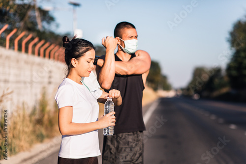 Asian couple with face mask working out and relaxing on street together. She drinking water and putting off mask and he stretching arms. Sport and coronavirus or covid-19 concept. 