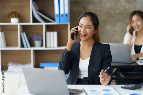 Happy young Asian business woman sitting smiling and talking on mobile phone in office.