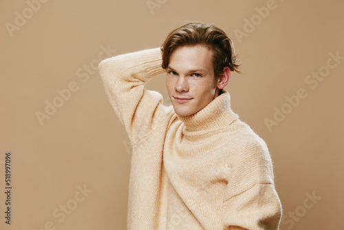 a cute pleasant relaxed young man in a beautiful fluffy sweater stands looking at the camera on a plain studio background and holds his hand behind his head. Horizontal photo with empty space © Tatiana