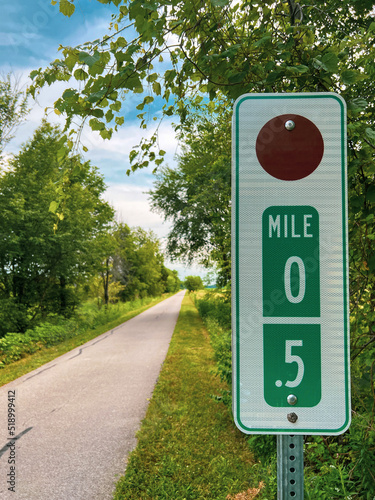 Mile Marker Trail Sign. Current distance covered is 0.5 miles. Lush green backdrop with leafy trees. Asphalt trail runs parallel to the sign. Cloudy blue skies above. photo