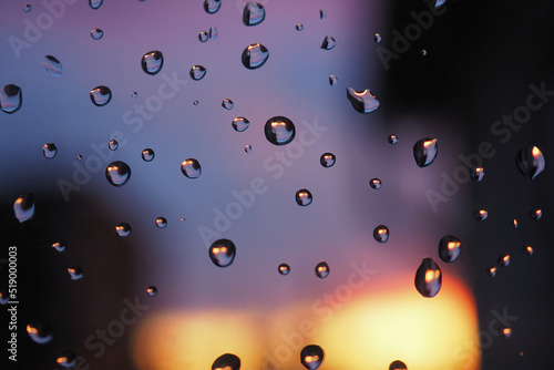 Raindrops on the window glass. In the background in a strong blur evening sunset. Dark blue purple and yellow backdrop. Abstract natural wallpaper. The rain is over. Macro