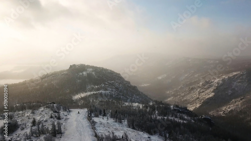 Aerial view of ski resort and snowy mountains covered by coniferous forest. Stock footage. White hill slope with ski tracks on cloudy sky background, sport and nature concept.