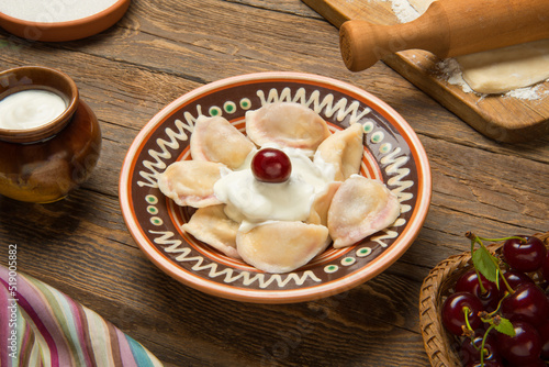 Traditional Ukrainian dumplings, vareniki with cherries in a ceramic painted bowl with ingredients on an old rural wooden table. photo