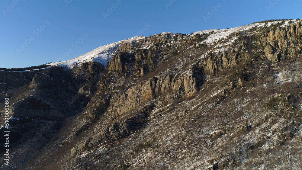 Aerial of snow covered hills with growing trees on the slopes on blue clear sky background. Shot. Camera moving away from beautiful rocks, natural background.