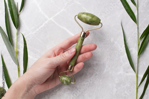 Fototapeta Naklejka Na Ścianę i Meble -  Woman hands holding roller for face massage on marble background with branch leaves. Anti age, lifting and toning treatment