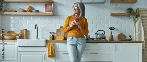 Thoughtful senior woman holding cup and smiling while standing at the domestic kitchen