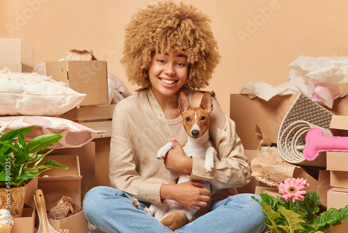 Indoor shot of happy young woman poses with favorite pet sits crossed legs around carton boxes moves to new apartment collects personal belongins isolated over brown background. Mortgage concept photo