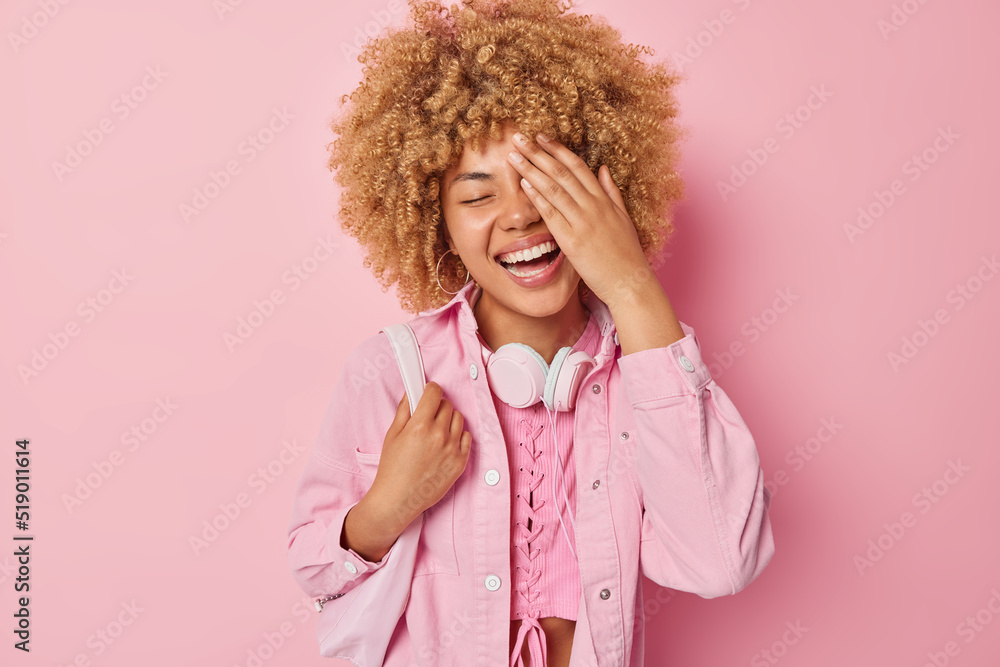 Indoor shot of positive woman with curly hair makes face palm giggles happily expresses happiness dressed casually carries bag isolated over pink background cannot stop laughing at something funny Indoor shot of positive woman with curly hair makes face palm giggles happily expresses happiness dressed casually carries bag isolated over pink background cannot stop laughing at something funny