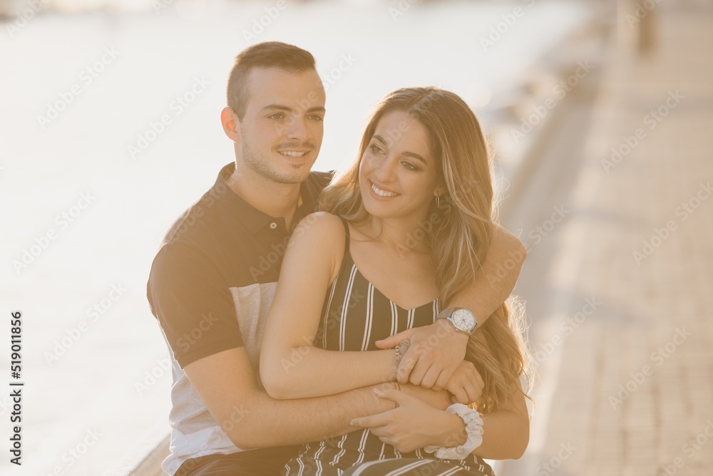 A couple of tourists are sitting near a water pool on a date at the sunset in Valencia. A guy is hugging his girlfriend. A girl is sitting between the legs of her boyfriend in the evening in Spain.