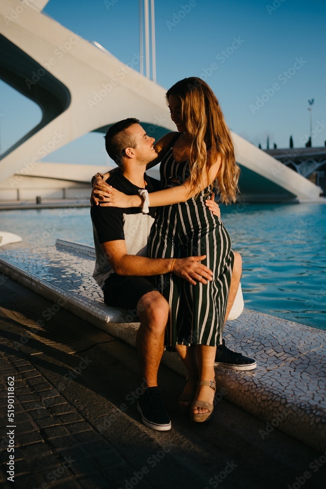 A couple of tourists are sitting in the modern urban space near a water pool on a date at the sunset in Valencia. A smiling girl is sitting between the legs of her boyfriend in the evening in Spain.