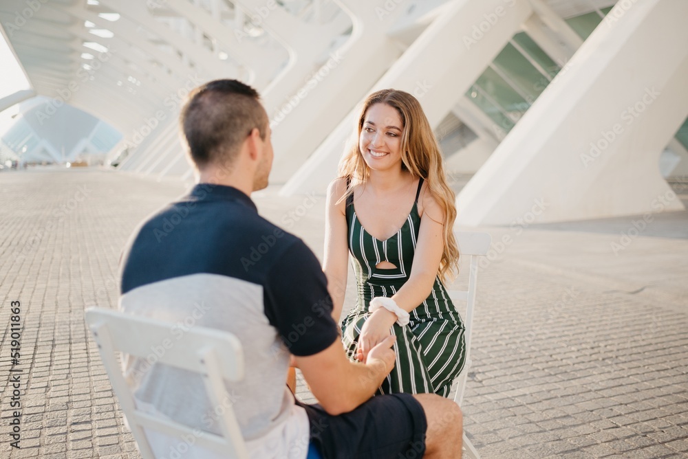 A couple of tourists are sitting in the modern urban space on a date at the sunset in Valencia. A smiling girl is holding hands with her boyfriend in the evening in Spain.