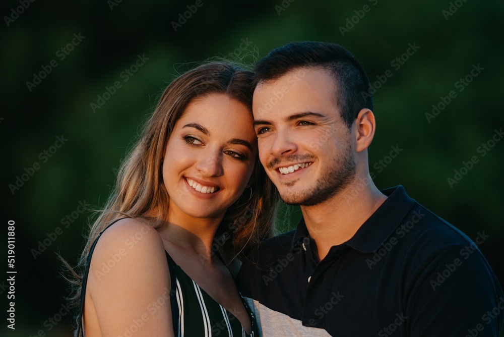 A couple of smiling tourists are having fun with green bushes in the background on a date at the sunset in Valencia. A guy is hugging his girlfriend in the evening in Spain