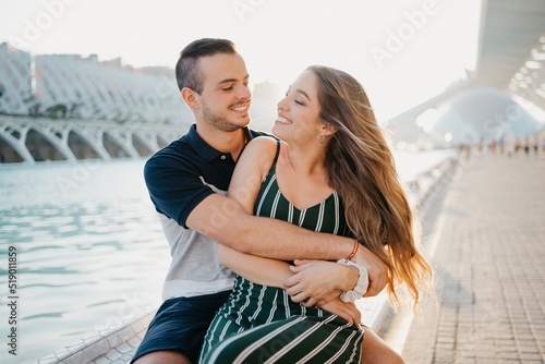 A smiling couple of tourists are having fun near a water pool on a date at the sunset in Valencia. A guy is hugging his girlfriend. A girl is sitting on the leg of her boyfriend in Spain
