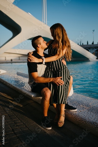Obraz na płótnie A couple of tourists are sitting in the modern urban space near a water pool on a date at the sunset in Valencia. A smiling girl is sitting between the legs of her boyfriend in the evening in Spain. w salonie