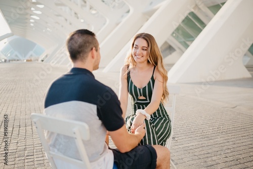A couple of tourists are sitting in the modern urban space on a date at the sunset in Valencia. A smiling girl is holding hands with her boyfriend in the evening in Spain.