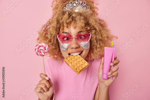 Indoor shot of funny curly haired woman holds lollipop waffle in mouth tube of toothpaste and toothbrush wears heart shaped sunglasses and t shirt isolated over pink background. Sugar addiction