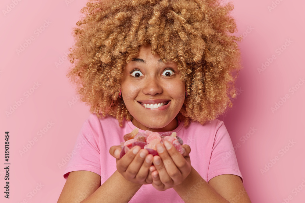 Surprised curly haired young woman holds jelly candies in hands bites
