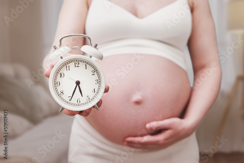 Alarm clock in the hands of a pregnant woman, home living room