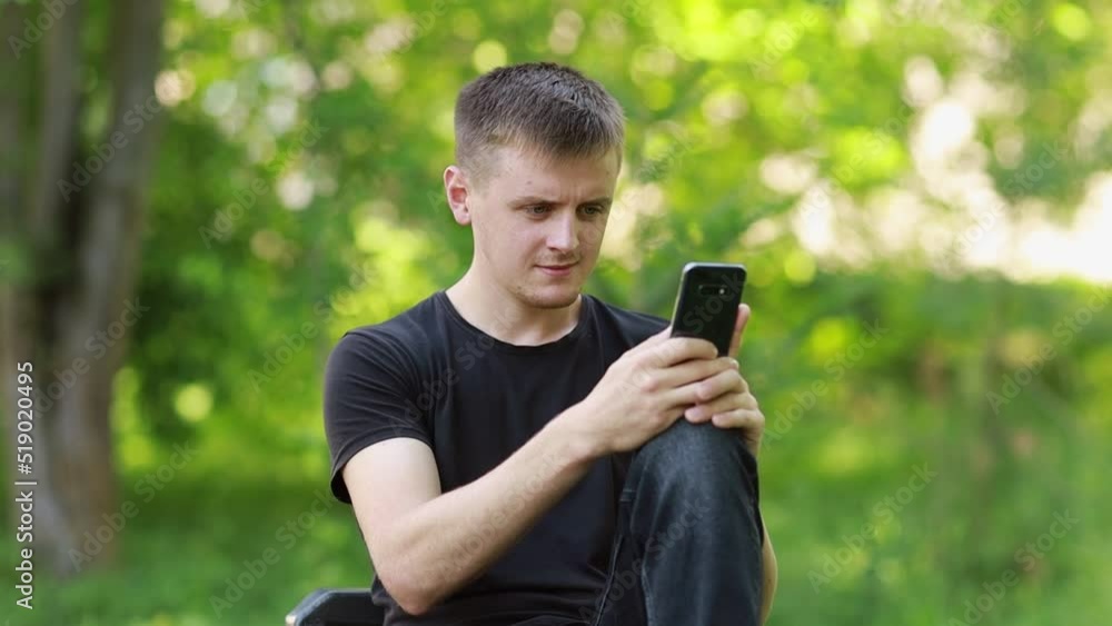A young freelancer works remotely from the park. The guy is viewing content from the phone, which is on his lap. A young freelancer works remotely from the park. The guy is viewing content from the phone, which is on his lap.