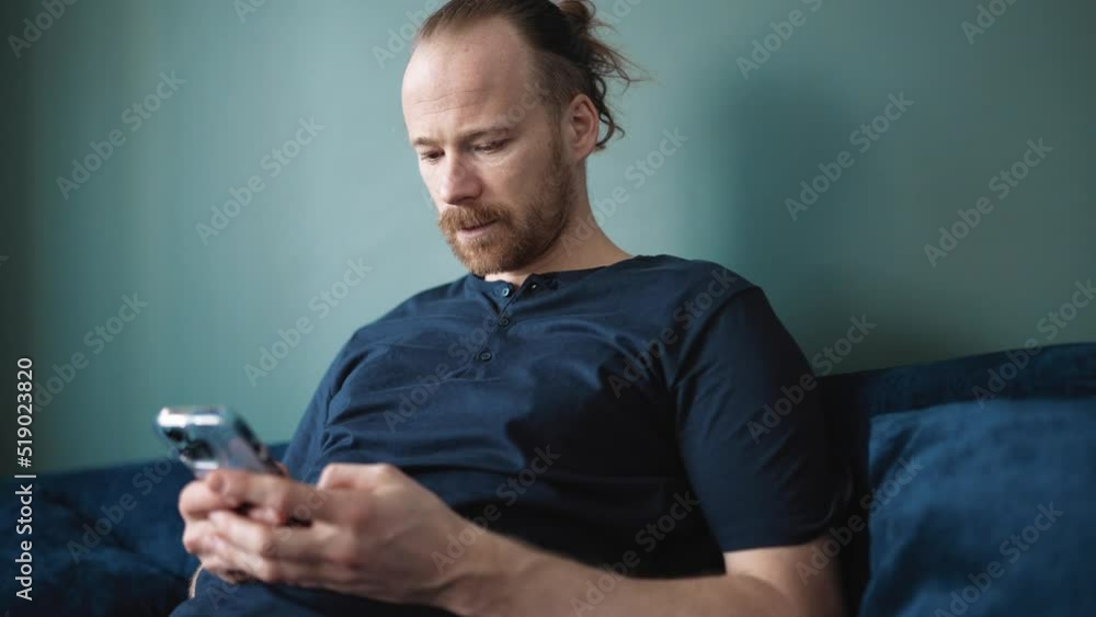 Laughing blond man wearing pajama texting on mobile while sitting on the sofa at home Laughing blond man wearing pajama texting on mobile while sitting on the sofa at home