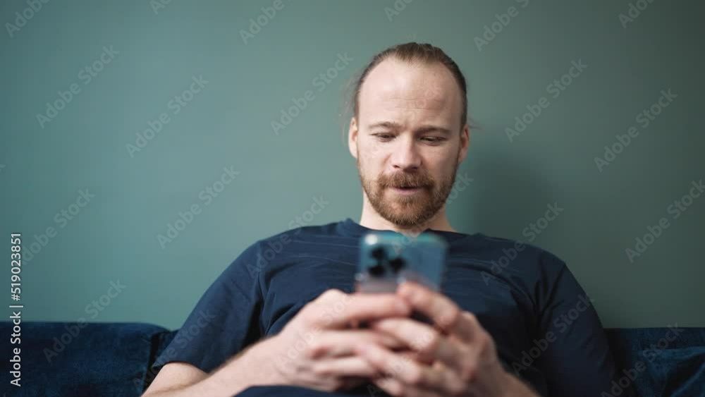 Pretty blond man wearing pajama texting on mobile while sitting on the sofa at home Pretty blond man wearing pajama texting on mobile while sitting on the sofa at home